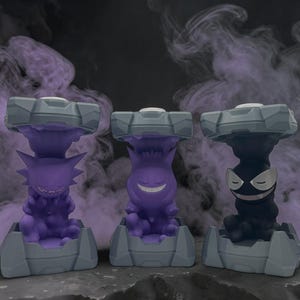 May include: Three stylized figures of Pokemon characters, each in a different color: purple, black, and purple. Each figure is set on a gray base with a matching gray top. The background is a dark gray with purple smoke.