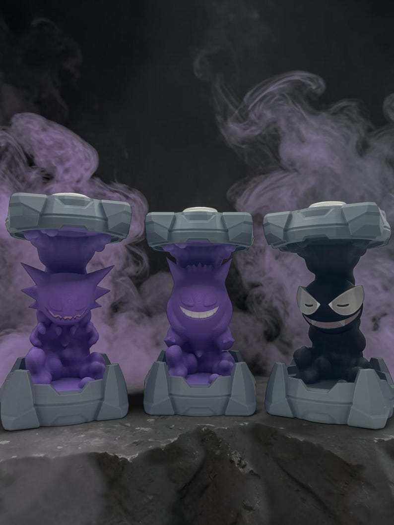 May include: Three stylized figures of Pokemon characters, each in a different color: purple, black, and purple. Each figure is set on a gray base with a matching gray top. The background is a dark gray with purple smoke.