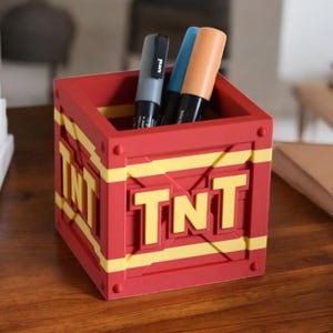 May include: A red desk organizer shaped like a TNT crate, with yellow accents and the letters "TNT" on the front. The organizer holds three pens: gray, blue, and orange. The desk organizer is made of plastic.