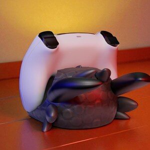 May include: A white and black gaming controller rests on top of a dark gray, textured object with rounded edges and protruding shapes. The controller has black accents and is positioned on a reddish-orange surface.