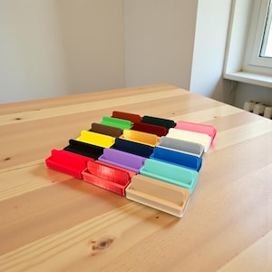 May include: A collection of colorful, rectangular organizers in various shades, including red, yellow, blue, and green, arranged on a light wooden surface. Each organizer has a raised back and a lower front, creating a tray-like design. The organizers are neatly aligned.