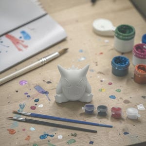 May include: A white, unpainted figurine of a cartoon character sits on a wooden surface, surrounded by paint pots and paintbrushes. Splatters of various colors of paint dot the surface, suggesting a creative project in progress. The figurine is the focal point.