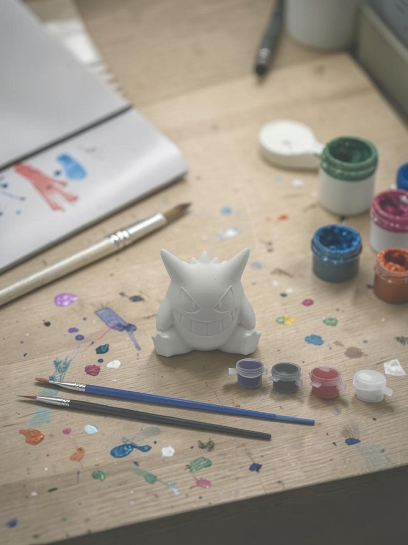 May include: A white, unpainted figurine of a cartoon character sits on a wooden surface, surrounded by paint pots and paintbrushes. Splatters of various colors of paint dot the surface, suggesting a creative project in progress. The figurine is the focal point.