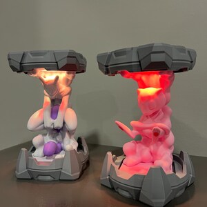 May include: Two gray and white 3D-printed lamps featuring a Pok&eacute;mon character design. One lamp glows with a warm white light, while the other emits a vibrant pink hue. The lamps have a geometric base and a cloud-like top.