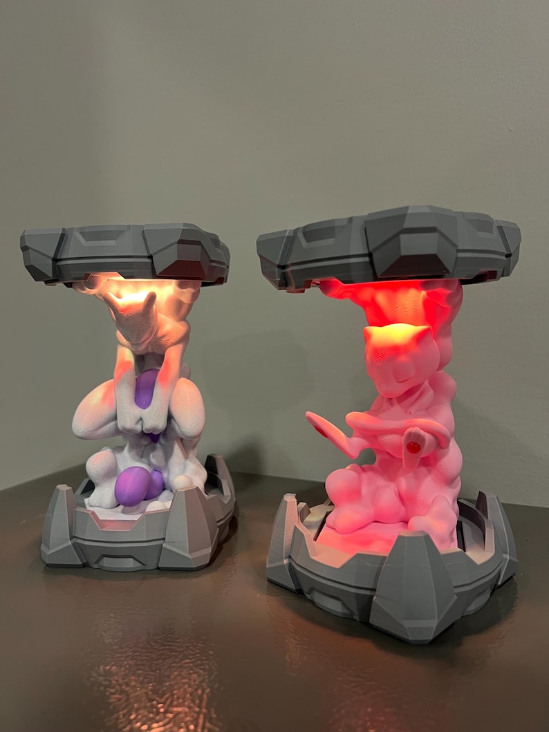 May include: Two gray and white 3D-printed lamps featuring a Pok&eacute;mon character design. One lamp glows with a warm white light, while the other emits a vibrant pink hue. The lamps have a geometric base and a cloud-like top.