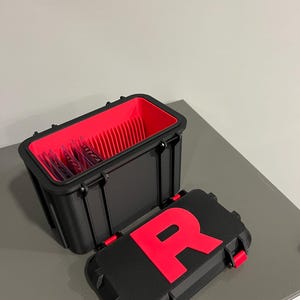 K&ouml;nnte beinhalten: Schwarze rechteckige Aufbewahrungsbox mit rotem Innenraum und passendem Deckel. Der Deckel hat ein gro&szlig;es rotes "R" und rote Verschl&uuml;sse. Die offene Box zeigt einen roten Innenraum mit vertikalen Trennw&auml;nden.