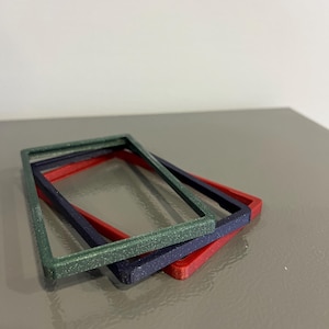 May include: Three rectangular frames in green, navy, and red are stacked on a gray surface. The green frame has a glittery texture. The frames are likely for crafts or decorative purposes.
