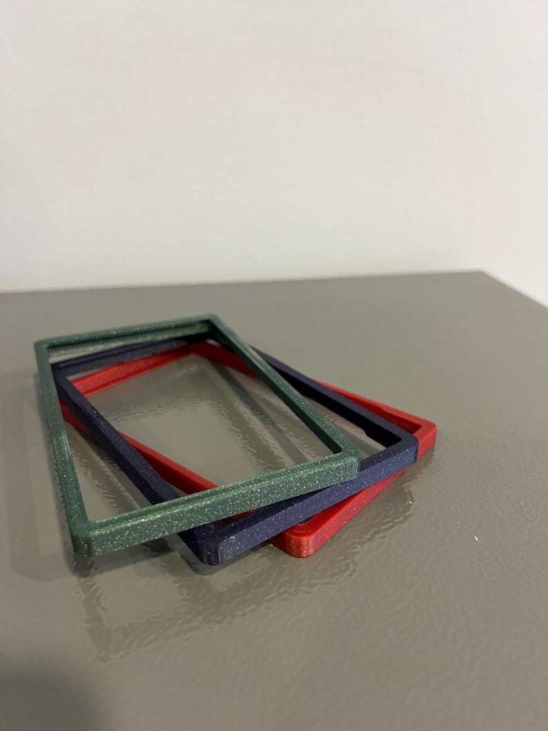 May include: Three rectangular frames in green, navy, and red are stacked on a gray surface. The green frame has a glittery texture. The frames are likely for crafts or decorative purposes.