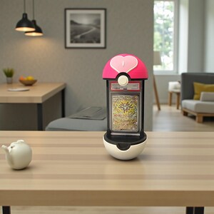 Puede incluir: Un expositor rosa y blanco con forma de Pok&eacute; Ball, que contiene una carta coleccionable calificada. El expositor tiene un dise&ntilde;o en forma de coraz&oacute;n en la parte superior. La tarjeta presenta una ilustraci&oacute;n colorida.
