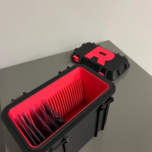 K&ouml;nnte beinhalten: Schwarzer rechteckiger Aufbewahrungsbeh&auml;lter mit rotem Innenraum und Trennw&auml;nden, der mehrere Kartenpackungen enth&auml;lt. Der Deckel mit einem gro&szlig;en roten "R" auf schwarzem Hintergrund liegt neben dem offenen Beh&auml;lter. Der Beh&auml;lter hat zwei schwarze Verschl&uuml;sse.
