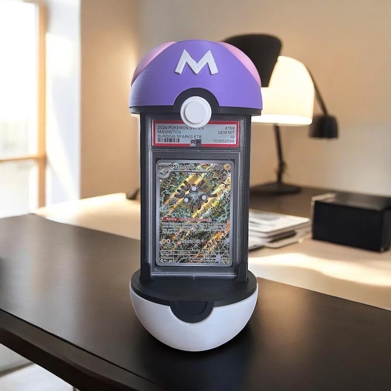 Op de afbeelding: Een Pok&eacute;mon-kaartdisplaystandaard in de vorm van een Pok&eacute; Ball, met een paarse bovenkant met een witte "M" en een witte knop. Het bevat een beoordeelde Pok&eacute;mon-kaart. De kaart is ingesloten in een doorzichtige plastic houder.