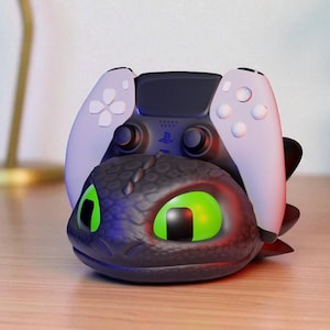May include: A dragon-themed PlayStation 5 controller stand in black and green. The stand, featuring a dragon head with glowing green eyes, holds a white and black controller. It sits on a wooden surface.