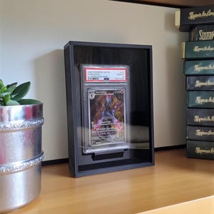 Può includere: Una cornice nera che espone una carta collezionabile Pokémon valutata. La carta presenta un'illustrazione di Ceruledge e il testo "2004 POKEMON SSP EN CERULEDGE ILLUSTRATION RARE GEM MT". La cornice è su una superficie di legno.