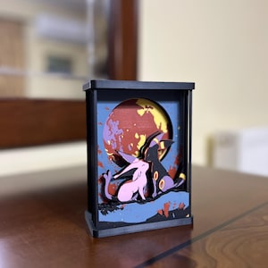 K&ouml;nnte beinhalten: Ein schwarzer Holzrahmen mit einem 3D-Diorama von zwei Pok&eacute;mon-Charakteren, einem rosa Espeon und einem lila Umbreon, die vor einem rot-gelb-blauen Mond stehen.
