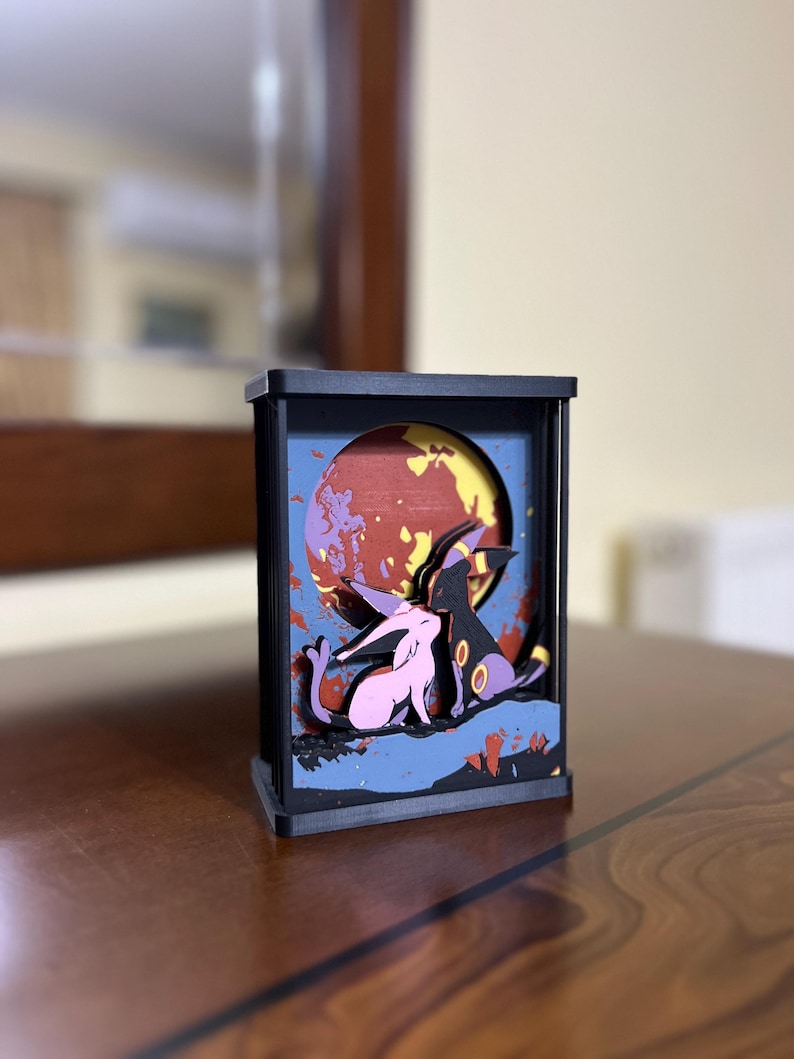 K&ouml;nnte beinhalten: Ein schwarzer Holzrahmen mit einem 3D-Diorama von zwei Pok&eacute;mon-Charakteren, einem rosa Espeon und einem lila Umbreon, die vor einem rot-gelb-blauen Mond stehen.