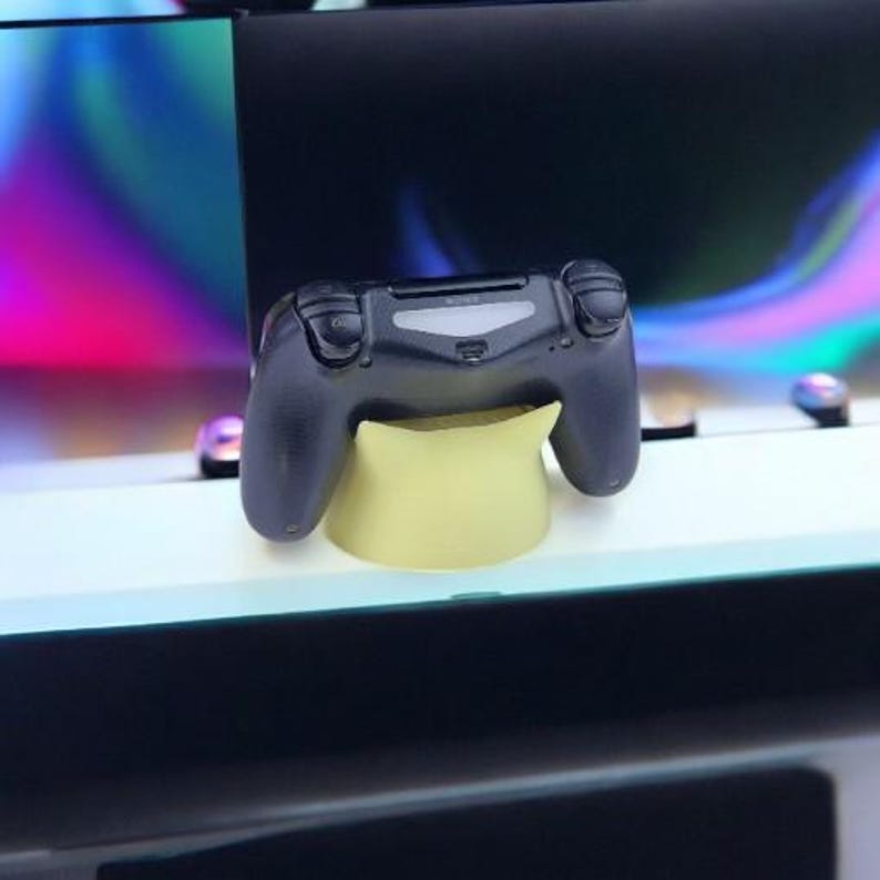 K&ouml;nnte beinhalten: Ein schwarzer drahtloser Gamecontroller mit einem gelben St&auml;nder. Der Controller hat Kn&ouml;pfe und ein Touchpad.