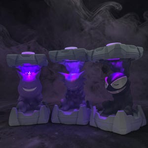 May include: Three stylized, gray and purple character lamps with glowing purple light. Each lamp features a different character design, with a smiling face, a spiky silhouette, and a closed-eye expression. The lamps are set against a dark, smoky background.