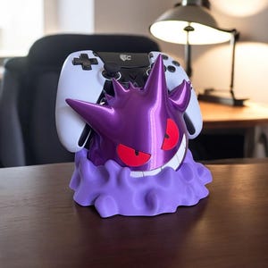 Può includere: Un supporto per controller viola a tema Gengar con occhi rossi e un sorriso bianco. Un controller bianco è appoggiato nel supporto. Il supporto è su una superficie di legno.