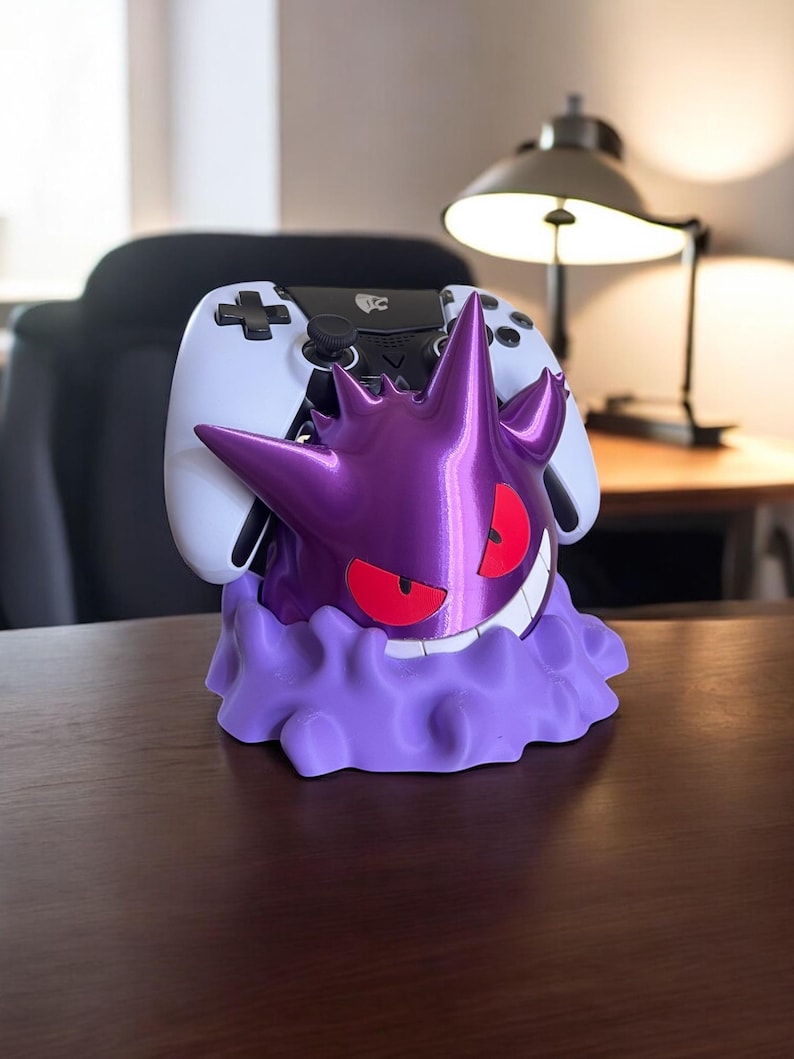 May include: A purple Gengar-themed controller holder with red eyes and a white smile. A white controller rests in the holder. The holder is on a wooden surface.