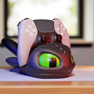 May include: A black and gray dragon-themed controller holder with a pink and black game controller. The dragon has large green eyes and is resting on a wooden surface. A small green plant and a gold lamp are in the background.