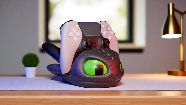 May include: A black and gray dragon-themed controller holder with a pink and black game controller. The dragon has large green eyes and is resting on a wooden surface. A small green plant and a gold lamp are in the background.