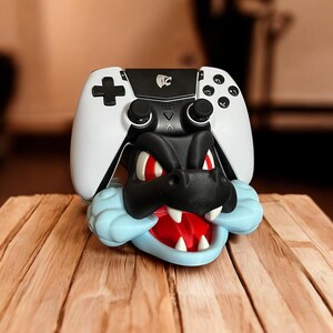 Op de afbeelding: Een wit en zwarte gamecontroller rust op een zwart en blauw draakachtig figuur. De draak heeft rode ogen en een rode mond met witte tanden. De controller heeft een kruisvormige knop en andere bedieningsknoppen.