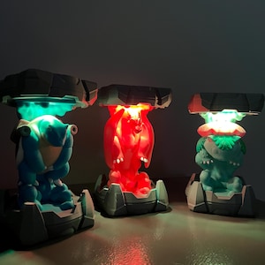 May include: Three illuminated character lamps in blue, red, and green. Each lamp features a unique character design with a rocky, gray base and a dark gray, stone-like top. The lamps emit a vibrant glow, creating a colorful display.