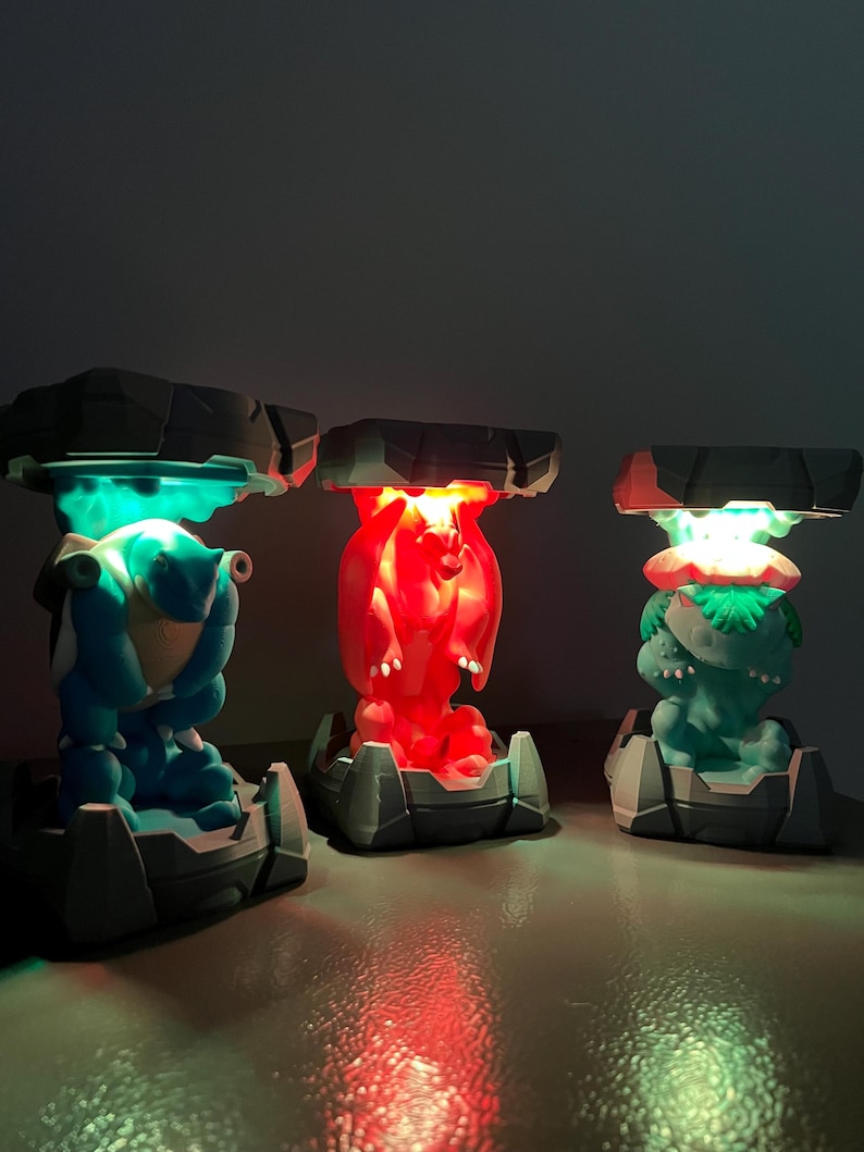 May include: Three illuminated character lamps in blue, red, and green. Each lamp features a unique character design with a rocky, gray base and a dark gray, stone-like top. The lamps emit a vibrant glow, creating a colorful display.