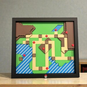 May include: A framed, layered artwork depicting a pixelated map with green, brown, and tan terrain. Blue water features with white wave patterns are visible. Red squares mark locations. A black frame surrounds the artwork, with a Pok&eacute; Ball detail.