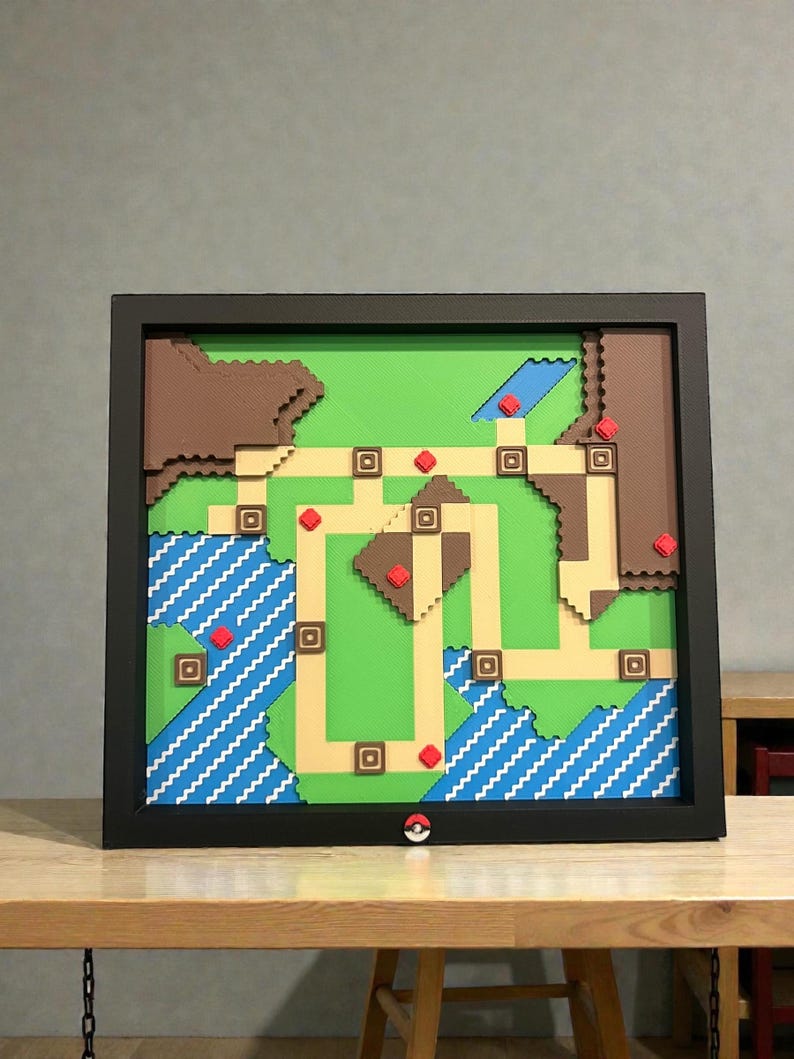 May include: A framed, layered artwork depicting a pixelated map with green, brown, and tan terrain. Blue water features with white wave patterns are visible. Red squares mark locations. A black frame surrounds the artwork, with a Pok&eacute; Ball detail.