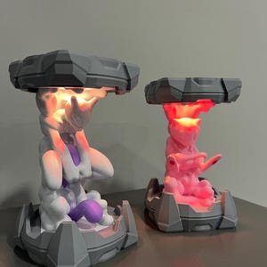 May include: Two 3D-printed lamps featuring a stylized creature design. One lamp is white and purple, the other is pink. Both have a gray, geometric top and base, with the creature in the center. The lamps emit a warm glow.