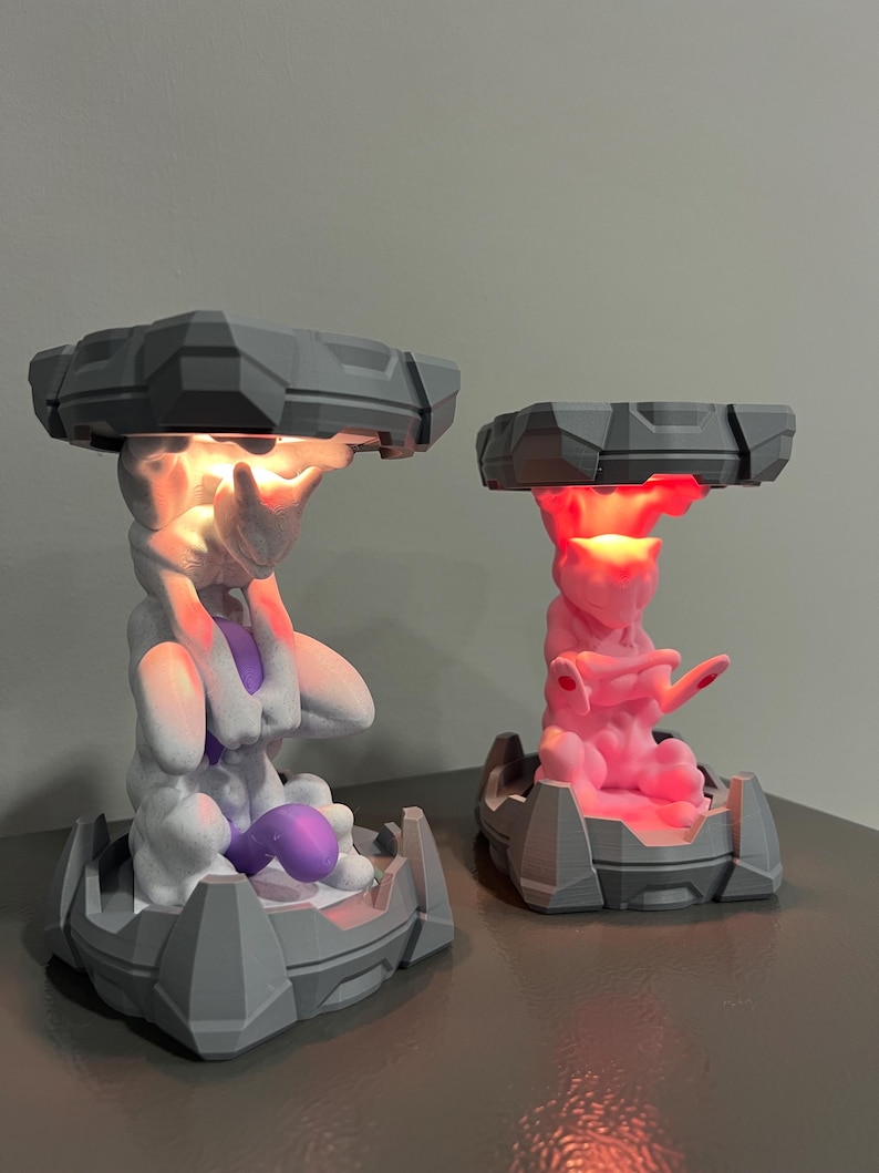 May include: Two 3D-printed lamps featuring a stylized creature design. One lamp is white and purple, the other is pink. Both have a gray, geometric top and base, with the creature in the center. The lamps emit a warm glow.