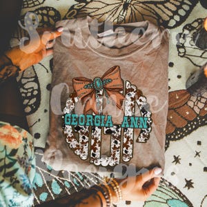 May include: A brown t-shirt with a white and brown cow print monogram design. The monogram is surrounded by a turquoise bow with a turquoise jewel in the center. The text "GEORGIA ANN" is written in turquoise letters above the monogram.
