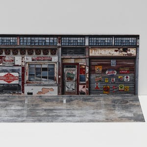 May include: A miniature diorama of an old auto repair shop with weathered facades. The shop features signs, windows, and a roll-up door. The scene is set on a gray concrete base, with a white background.