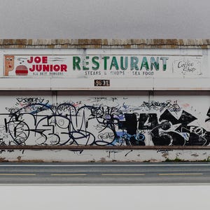 Peut inclure: Maquette miniature d'une façade de restaurant. Le bâtiment est blanc avec un toit marron et une scène de rue peinte à la base. L'enseigne du restaurant indique "Joe Junior Restaurant" et "Steaks & Chops Sea Food". La façade est recouverte de graffitis.