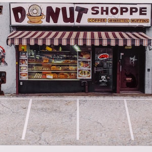 Puede incluir: Modelo en miniatura de una tienda de donuts con un toldo a rayas rojas y blancas. El letrero dice "DONUT SHOPPE" con un gráfico de donut y taza de café. La tienda tiene una exhibición de productos horneados y un estacionamiento.