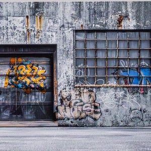 Puede incluir: Un muro de hormigón desgastado con una puerta de garaje y una ventana, ambas con marcos metálicos. La puerta del garaje presenta arte de graffiti colorido con una flecha. La ventana tiene un patrón de cuadrícula. La pared está cubierta de graffiti y muestra signos de edad y óxido.