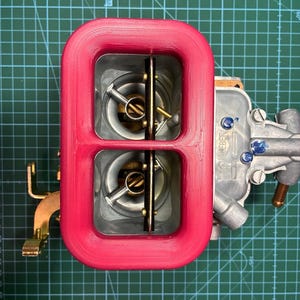 Puede incluir: Un carburador rojo con dos cámaras y componentes internos metálicos. El carburador tiene un cuerpo plateado con la palabra "Weber" impresa. El fondo es una esterilla de corte verde con líneas de cuadrícula.