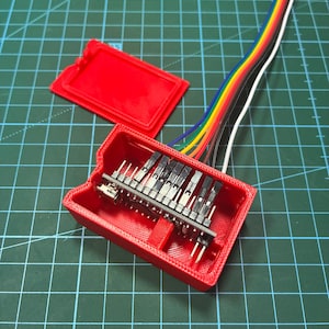 May include: A bright red 3D-printed plastic enclosure with a matching lid, containing a small electronic microcontroller board with multiple black pin headers. A multi-colored ribbon cable with individual wires in red, orange, yellow, green, blue, and white extends from the board.