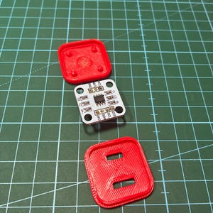 May include: A square white electronic circuit board with a central black integrated chip and labeled pins, positioned between two red 3D-printed plastic covers. The top cover has four small circular protrusions, and the bottom cover has two rectangular slots.