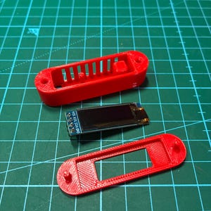 以下が含まれることがあります： 電子部品用の赤い3Dプリント製プラスチックケース。通気スロット付きのトップカバーと長方形の切り抜きがあるボトムプレートで構成され、ピン付きの小さな長方形のOLEDディスプレイモジュールがその間に配置されています。