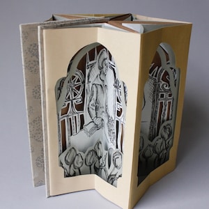 May include: A pop-up book with a beige cover and a cut-out window revealing a black and white illustration of a person sitting in a garden with tulips. The book is folded into a star shape.