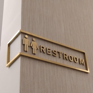 May include: A gold-colored restroom sign with male and female figures and the word "RESTROOM" in capital letters. The sign is mounted on a textured, light-colored wall and framed with a gold border.