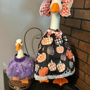 Pu&ograve; includere: Due statuette decorative a forma di oca. Una indossa un vestito nero con motivi di zucche arancioni e un cappello abbinato. L'altra indossa un tut&ugrave; viola. Entrambe hanno becchi e zampe arancioni. Gli abiti sono rifiniti con pizzo.