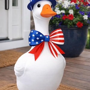 Pu&ograve; includere: Una statua di un'oca bianca con cappello e papillon patriottici. Il cappello &egrave; rosso, bianco e blu, e il papillon presenta una sezione blu con stelle bianche e una sezione a righe rosse e bianche. L'oca ha zampe e becco arancioni.