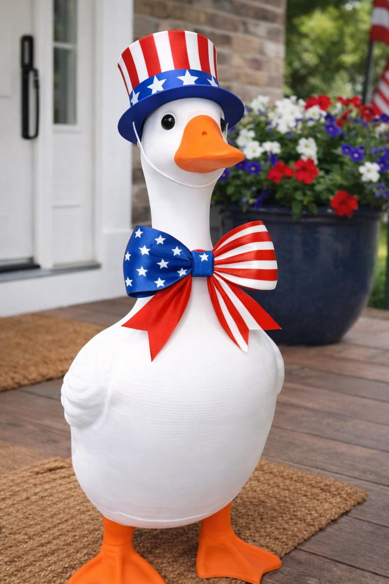 Pu&ograve; includere: Una statua di un'oca bianca con cappello e papillon patriottici. Il cappello &egrave; rosso, bianco e blu, e il papillon presenta una sezione blu con stelle bianche e una sezione a righe rosse e bianche. L'oca ha zampe e becco arancioni.
