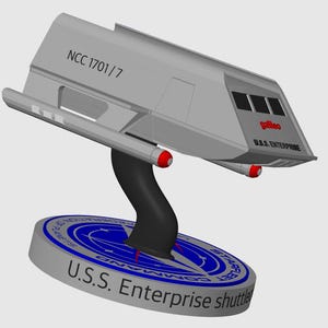 May include: A gray model of a Star Trek shuttlecraft, the U.S.S. Enterprise shuttle, with the registration number NCC 1701/7 and the name "Galileo" on the side. The shuttle is mounted on a black stand with a blue Starfleet Command emblem on a gray base.