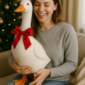 Pu&ograve; includere: Un'oca bianca di peluche di grandi dimensioni con becco e zampe arancioni e un papillon rosso. L'oca &egrave; tenuta da una persona che indossa una felpa grigia e jeans blu. Lo sfondo include un albero di Natale.