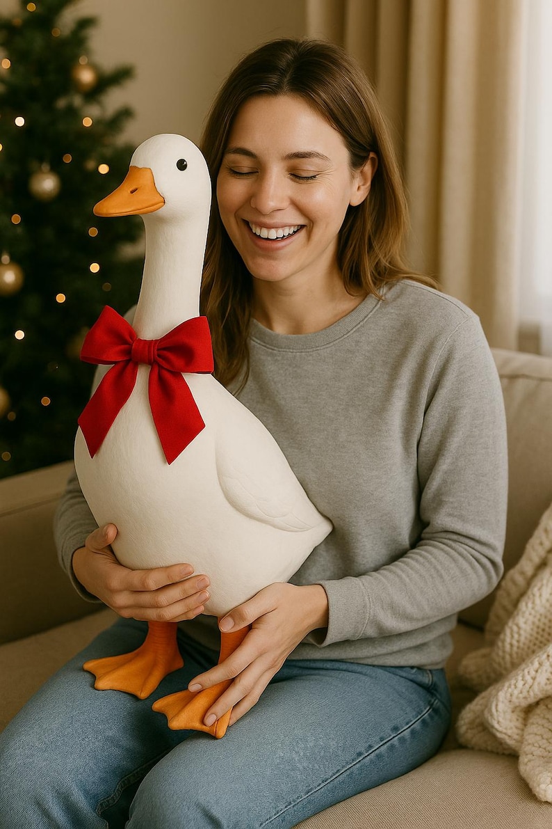 Pu&ograve; includere: Un'oca bianca di peluche di grandi dimensioni con becco e zampe arancioni e un papillon rosso. L'oca &egrave; tenuta da una persona che indossa una felpa grigia e jeans blu. Lo sfondo include un albero di Natale.