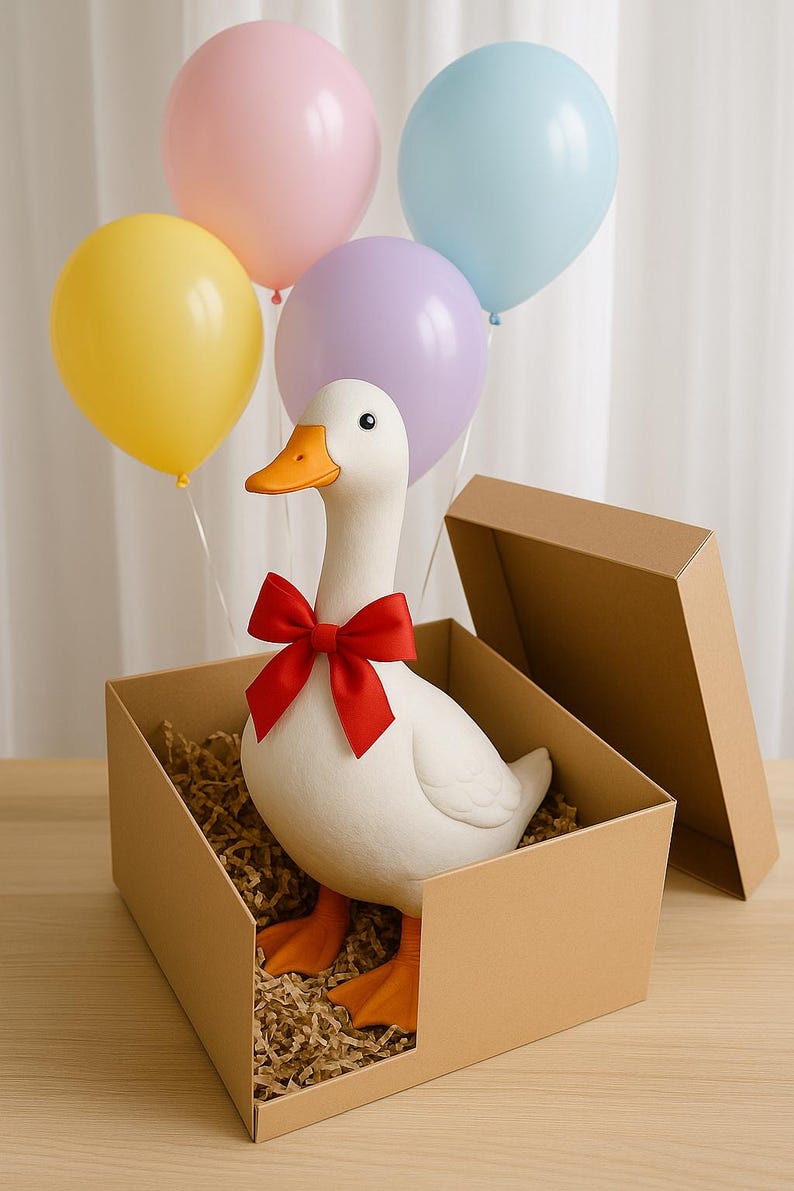 Pu&ograve; includere: Un'anatra di ceramica bianca con becco e zampe arancioni, con un fiocco rosso, &egrave; seduta in una scatola di cartone piena di imbottitura di carta. Palloncini pastello gialli, rosa, viola e blu fluttuano sopra. Un regalo o un oggetto decorativo stravagante.